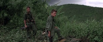 Movie still from “Casualties of War” (1989), directed by Brian De Palma – A man with a gun standing next to another man in the woods; Medium shot, Low angle
