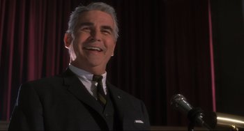 Movie still from “Catch Me If You Can” (2002), directed by Steven Spielberg – An older man wearing a suit and tie with a microphone in front of him; Close Up shot, Low angle