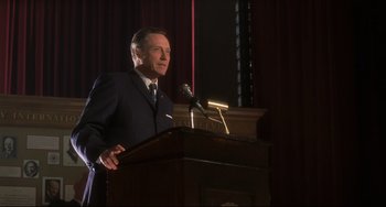 Movie still from “Catch Me If You Can” (2002), directed by Steven Spielberg – A man standing at a podium with a microphone; Medium shot, Low angle