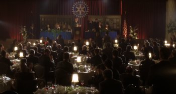 Movie still from “Catch Me If You Can” (2002), directed by Steven Spielberg – A group of people sitting at tables in front of a microphone; Extreme Wide shot, High angle
