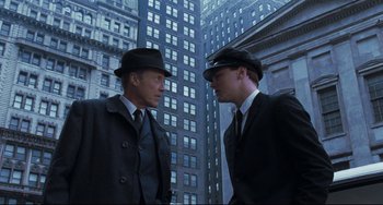 Movie still from “Catch Me If You Can” (2002), directed by Steven Spielberg – Two men in suits and hats standing next to each other in front of tall buildings; Medium shot, Low angle