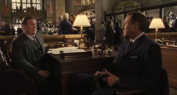 Movie still from “Catch Me If You Can” (2002), directed by Steven Spielberg – A man sitting at a desk in a suit and tie in front of another man; Medium shot, Over the shoulder angle