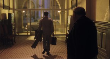 Movie still from “Catch Me If You Can” (2002), directed by Steven Spielberg – A man in a suit carrying two suitcases in front of a building; Wide shot, Over the shoulder angle