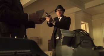 Movie still from “Catch Me If You Can” (2002), directed by Steven Spielberg – A man in a black suit and a black hat holding a gun; Medium shot, Low angle