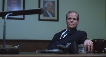 Movie still from “Catch Me If You Can” (2002), directed by Steven Spielberg – A man sitting at a table in front of a picture of george washington on the wall; Medium shot, Low angle