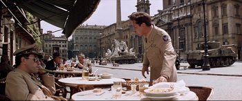 Movie still from “Catch-22” (1970), directed by Mike Nichols – A man standing at a table with plates of food on top of it; Wide shot, High angle