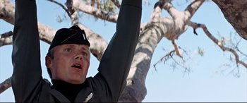 Movie still from “Catch-22” (1970), directed by Mike Nichols – A man in a black jacket and a black hat is in front of a large tree; Close Up shot, Low angle