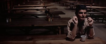 Movie still from “Catch-22” (1970), directed by Mike Nichols – A man sitting on top of a wooden bench next to wooden tables; Medium shot, High angle