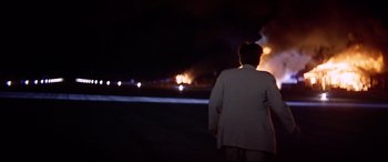 Movie still from “Catch-22” (1970), directed by Mike Nichols – A man in a white suit standing in front of a fire; Extreme Wide shot, Over the shoulder angle