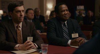 Movie still from “Cedar Rapids” (2011), directed by Miguel Arteta – Two men sitting at a table in front of other people; Medium shot, Over the shoulder angle