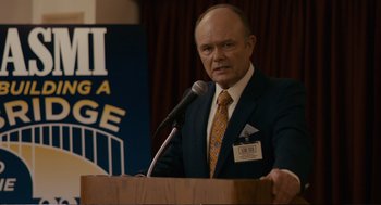 Movie still from “Cedar Rapids” (2011), directed by Miguel Arteta – A man in a business suit is speaking at a podium; Medium shot, Over the shoulder angle