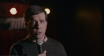 Movie still from “Cedar Rapids” (2011), directed by Miguel Arteta – A man holding a microphone in front of a dark background; Close Up shot, Over the shoulder angle