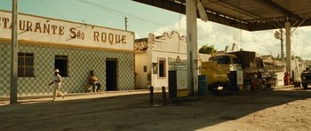 Movie still from “Central Station” (1998), directed by Walter Salles – A gas station with people sitting on the side of the road; Extreme Wide shot, Over the shoulder angle