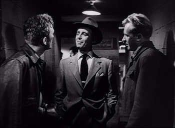 Movie still from “Champion” (1949), directed by Mark Robson – A man in a suit and hat standing next to two other men; Medium shot, Over the shoulder angle