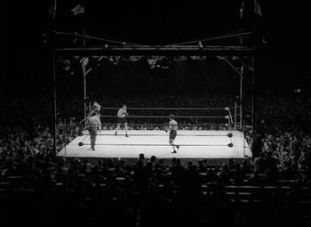 Movie still from “Champion” (1949), directed by Mark Robson – A boxing ring with three men on it and a crowd watching; Extreme Wide shot, High angle