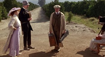 Movie still from “Chaplin” (1992), directed by Richard Attenborough – A man in a trench coat and hat holding a suitcase; Wide shot, Low angle