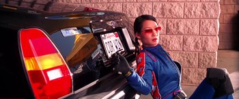 Movie still from “Charlie's Angels” (2000), directed by McG – A woman holding a license plate next to a parked car; Medium shot, Low angle