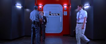 Movie still from “Charlie's Angels” (2000), directed by McG – A man in a suit talks to a police officer in front of a door; Wide shot, Over the shoulder angle