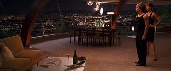 Movie still from “Charlie's Angels” (2000), directed by McG – A table with a wine bottle and a plate of food on top of it in front of a dining room table with a city view; Extreme Wide shot, High angle