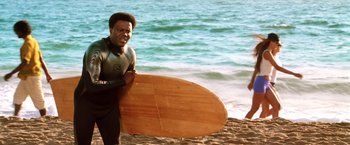 Movie still from “Charlie's Angels: Full Throttle” (2003), directed by McG – A man holding a surfboard while standing on the beach; Medium shot, Over the shoulder angle