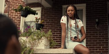 Movie still from “Charm City Kings” (2020), directed by Angel Manuel Soto – A woman sitting on a ledge holding a camera; Medium shot, Over the shoulder angle