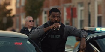 Movie still from “Charm City Kings” (2020), directed by Angel Manuel Soto – A police officer talking on a phone while standing next to a car; Medium shot, Over the shoulder angle