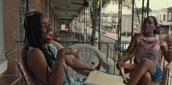 Movie still from “Charm City Kings” (2020), directed by Angel Manuel Soto – A woman sitting at a table outside on a porch; Medium shot, Over the shoulder angle
