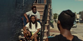 Movie still from “Charm City Kings” (2020), directed by Angel Manuel Soto – A couple of people that are sitting on some steps; Medium shot, High angle