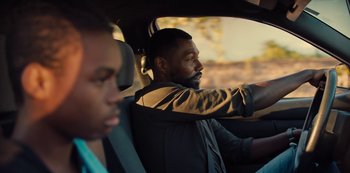 Movie still from “Charm City Kings” (2020), directed by Angel Manuel Soto – A man driving a car with another man sitting in the passenger seat; Close Up shot, Over the shoulder angle
