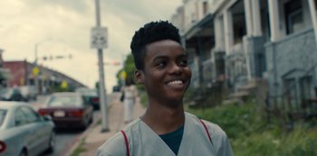 Movie still from “Charm City Kings” (2020), directed by Angel Manuel Soto – A young man smiling while standing on the side of the street; Close Up shot, Over the shoulder angle