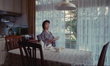Movie still from “Chizuko's Younger Sister” (1991), directed by Nobuhiko Ôbayashi – A woman sitting at a table reading a magazine; Medium shot, Low angle