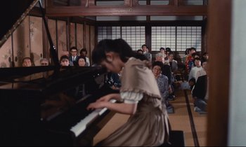 Movie still from “Chizuko's Younger Sister” (1991), directed by Nobuhiko Ôbayashi – A woman in a brown dress playing a piano in front of an audience; Wide shot, High angle