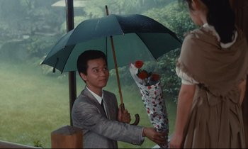 Movie still from “Chizuko's Younger Sister” (1991), directed by Nobuhiko Ôbayashi – A man holding an umbrella with a bouquet of flowers; Medium shot, Over the shoulder angle