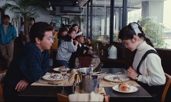 Movie still from “Chizuko's Younger Sister” (1991), directed by Nobuhiko Ôbayashi – A group of people sitting at a table with plates of food on it; Medium shot, Over the shoulder angle