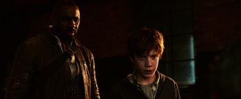 Movie still from “The Dark Tower” (2017), directed by Nikolaj Arcel – A man standing next to a young boy in a dark room; Medium shot, Over the shoulder angle