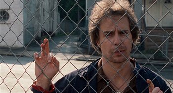 Movie still from “Choke” (2008), directed by Clark Gregg – A man holding up a peace sign behind a chain link fence; Close Up shot, Low angle