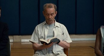 Movie still from “Choke” (2008), directed by Clark Gregg – An older man sitting down reading a book; Close Up shot, Low angle