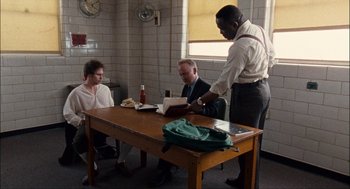 Movie still from “Choke” (2008), directed by Clark Gregg – Three men sitting at a table looking at a book; Wide shot, High angle