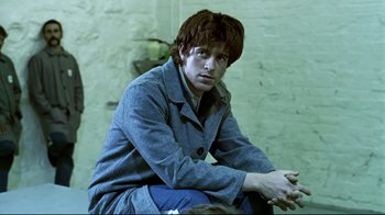 Movie still from “Chopper” (2000), directed by Andrew Dominik – A man sitting on the ground wearing a coat; Medium shot, Over the shoulder angle