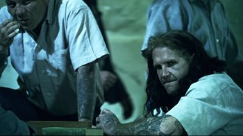 Movie still from “Chopper” (2000), directed by Andrew Dominik – A man with long hair and tattoos sitting at a table; Medium shot, Over the shoulder angle