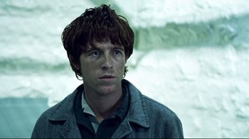 Movie still from “Chopper” (2000), directed by Andrew Dominik – A young man with long brown hair wearing a gray jacket; Close Up shot, Low angle