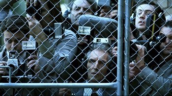 Movie still from “Chopper” (2000), directed by Andrew Dominik – A group of people standing behind a chain link fence; Medium shot, High angle
