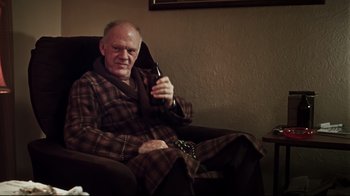 Movie still from “Chopper” (2000), directed by Andrew Dominik – An older man sitting in a chair holding a cell phone; Medium shot, Over the shoulder angle