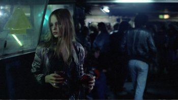 Movie still from “Christiane F.” (1981), directed by Uli Edel – A woman standing in a crowd holding a drink; Medium shot, Over the shoulder angle
