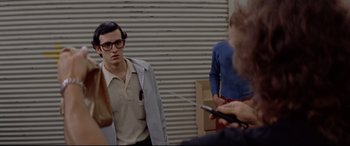 Movie still from “Christine” (1983), directed by John Carpenter – A young man wearing glasses is standing next to a woman; Medium shot, Over the shoulder angle