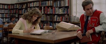 Movie still from “Christine” (1983), directed by John Carpenter – A woman sitting at a table reading a book in front of a bookcase; Medium shot, High angle