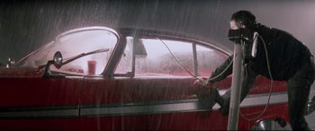 Movie still from “Christine” (1983), directed by John Carpenter – A person holding a hose in a red car in the pouring rain; Wide shot, Low angle
