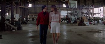 Movie still from “Christine” (1983), directed by John Carpenter – A man and a woman holding hands while walking in an airplane hangar; Wide shot, Low angle