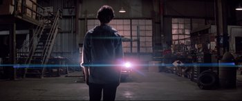 Movie still from “Christine” (1983), directed by John Carpenter – A man standing in a warehouse with a light shining on him; Wide shot, Over the shoulder angle