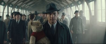 Movie still from “Christopher Robin” (2018), directed by Marc Forster – A man holding a teddy bear while standing next to a crowd; Medium shot, Low angle